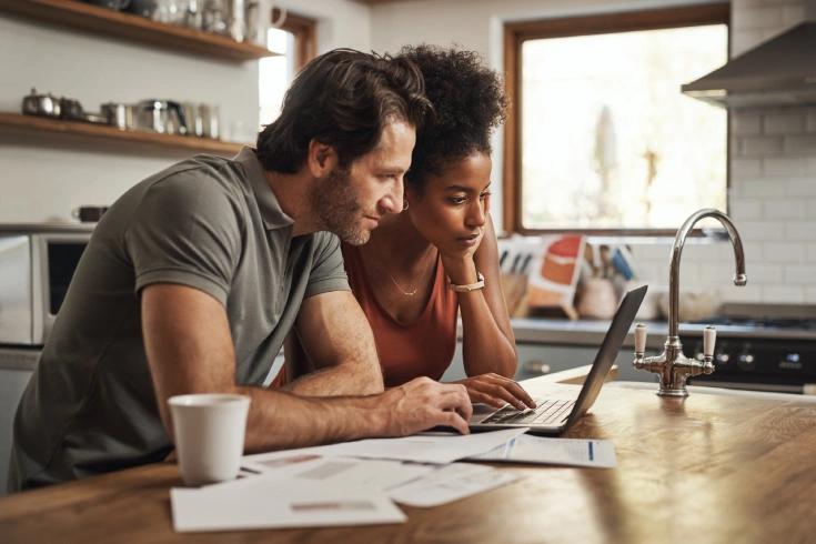 Interracial couple looking at a laptop in a kitchen with documents for budget, savings and paying bills