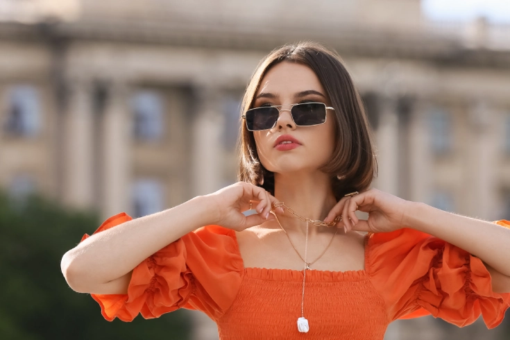 Woman with stylish jewellery and sunglasses touching necklace on city street