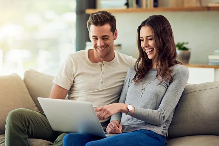A young married couple sit on the couch at home and review life insurance options on a laptop