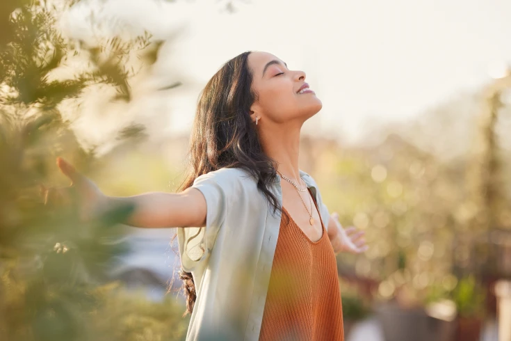A young woman stretches her arms out as she enjoys being outside in nature and the sunshine