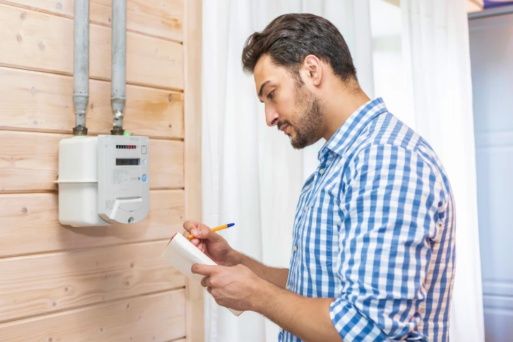 Middle-aged man in a checkered shirt, reading a meter on the wall with a pad and pen in hand