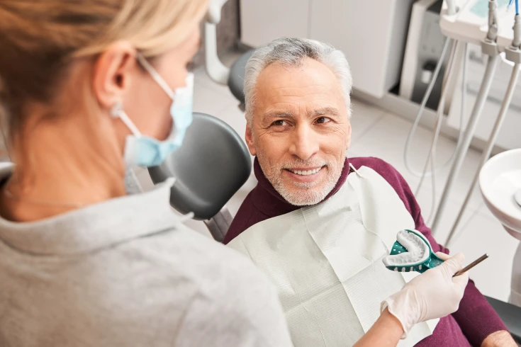 An older man smiles up at a dentist as she shows him the dental impression mould used to shape his dentures
