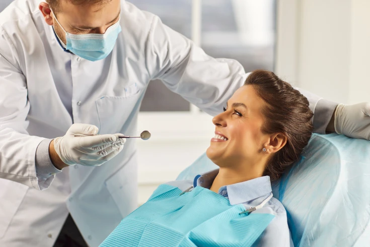 A woman leans back in a dental chair smiling as a dentist prepares to use a small mirror to check her teeth