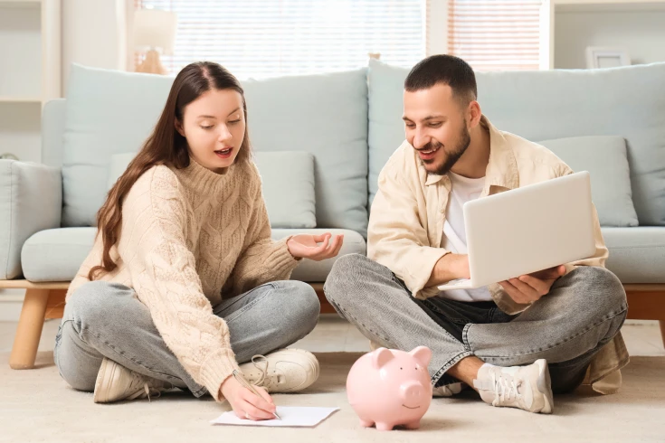 Man and woman sitting on the floor with the man holding a laptop, woman writing on paper, and a piggybank in front of them