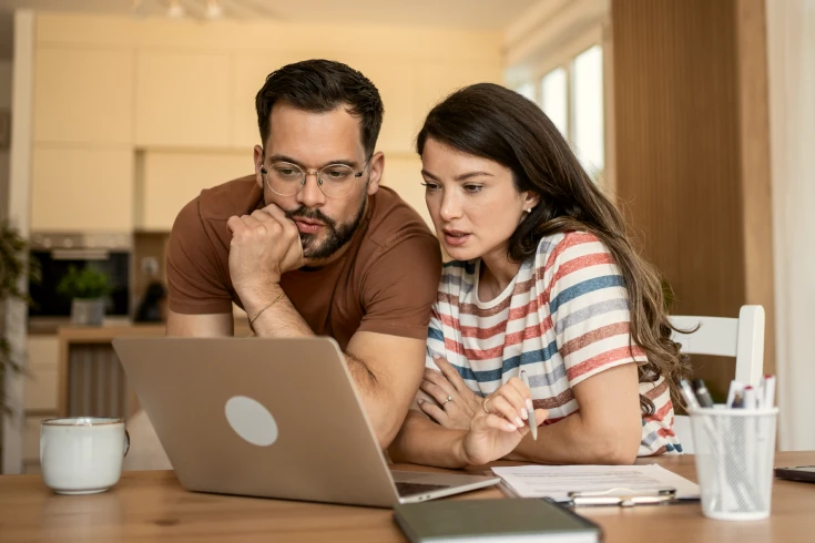Couple looking at laptop with woman holding a pen with paperwork in front of her