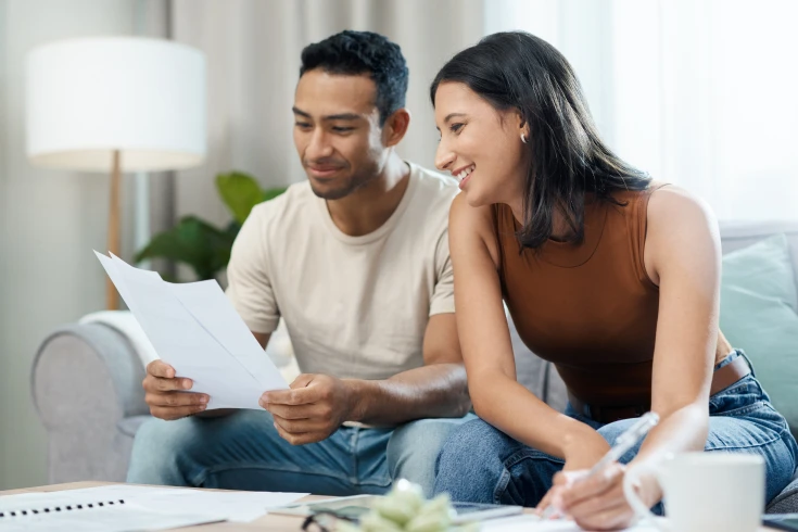 Couple sitting on couch, man is holding their average electricity bill, woman is writing on the coffee table
