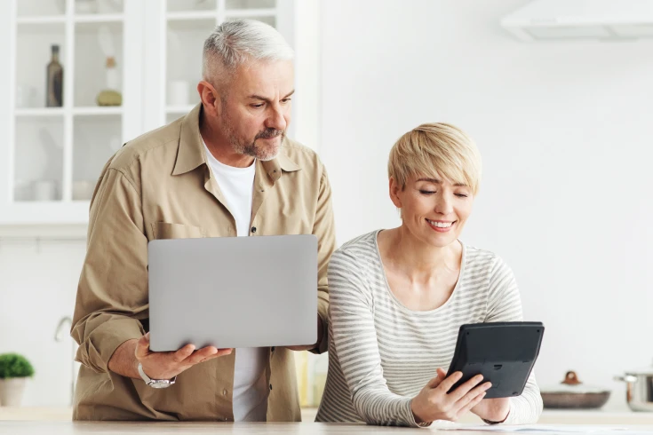 Couple standing in kitchen, a man with a laptop in his hand, a woman with a black tablet in her hand.
