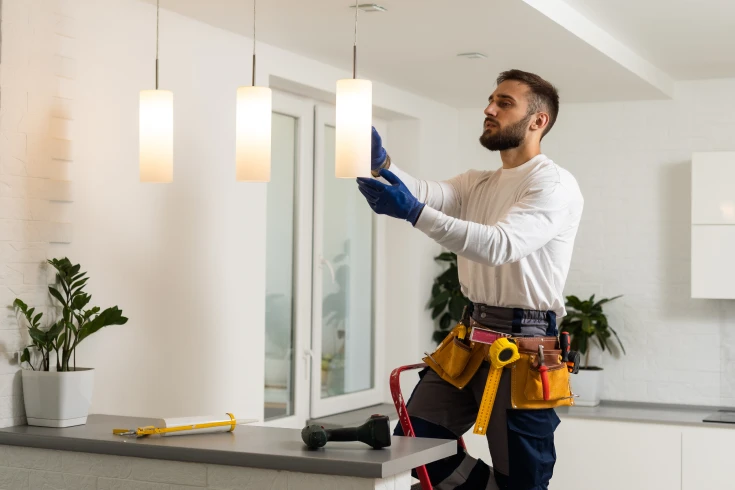 Man with tool belt on, standing on a ladder inside a house, changing hanging decorative lights.