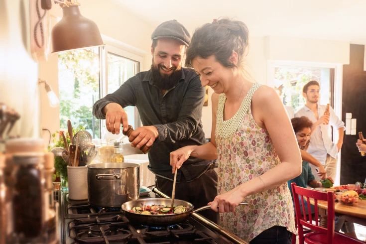 Man and woman both cooking at an oven cooktop with people in the background at a table