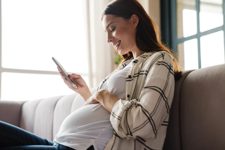 Pregnant woman sitting on a couch, smiling while using her phone.