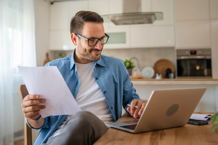 Man wearing glasses in the kitchen holding a sheet of paper on one hand while typing on the laptop with the other