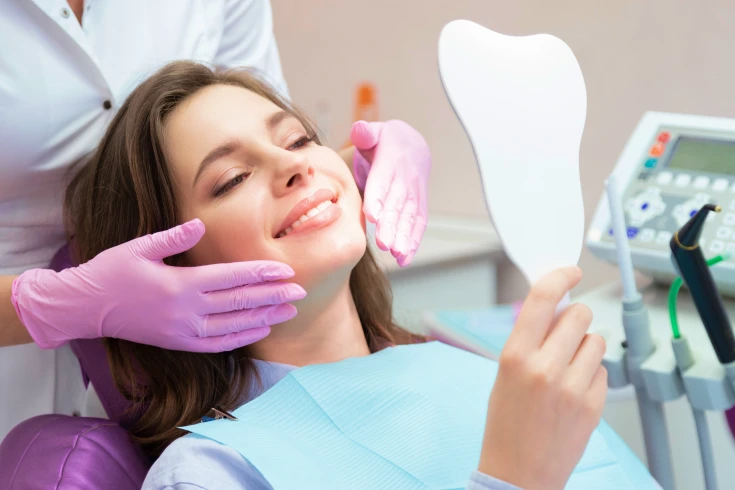 Woman in the dentist’s chair looking at herself in the mirror, smiling, with the dentist behind her wearing gloves