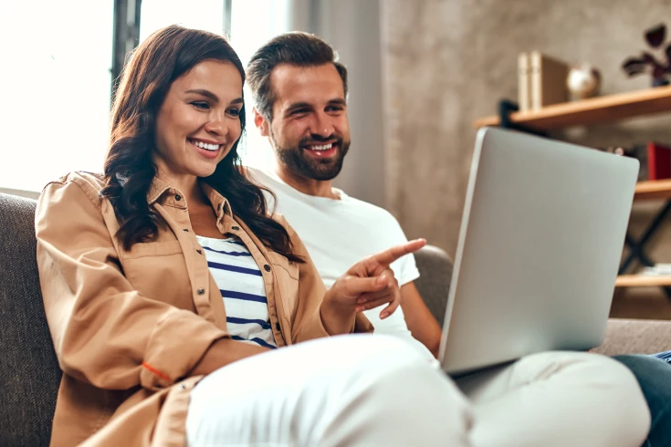 A couple sit on a couch, look at a laptop and smile