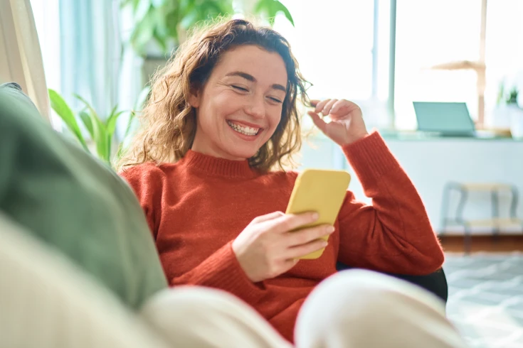 A woman reclines on a couch and smiles into her smartphone