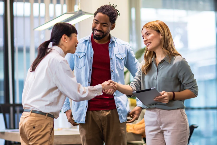 Two women and one man standing in an office environment, smiling and chatting as they greet each other.