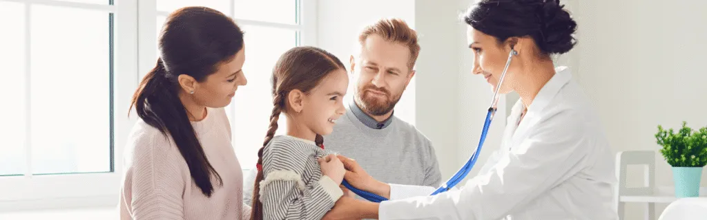 A female doctor checks the heart of a small girl with her mother and father present.
