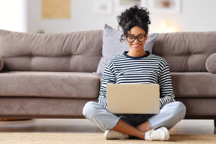 A woman sits on her living room floor and types on her laptop