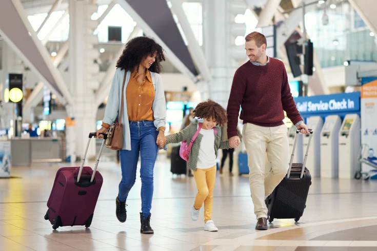 Family of three walking through airport, mother and father both have suitcases, holding hands of the young daughter in the middle