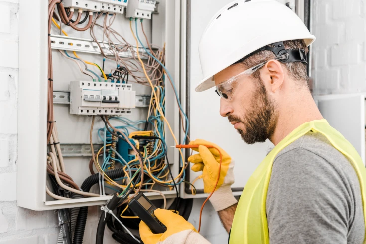 A tradesman wearing a hard hat, safety vest, and gloves working on an electrical panel.