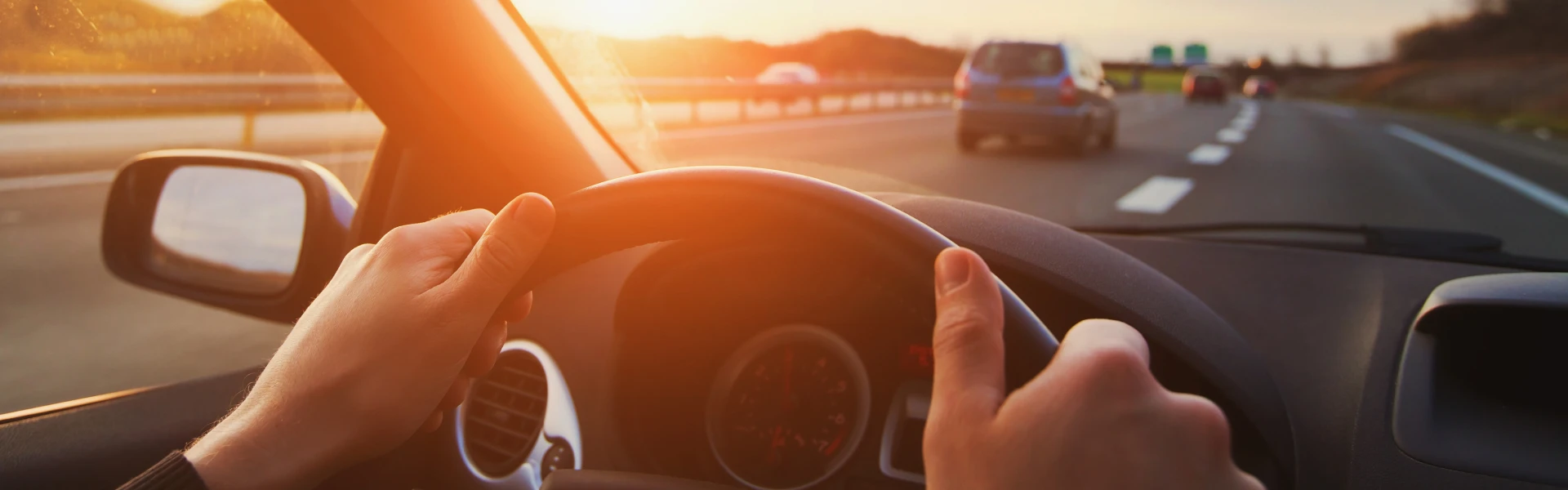 A driver has their hands on a steering wheel as they drive on a freeway at sundown.