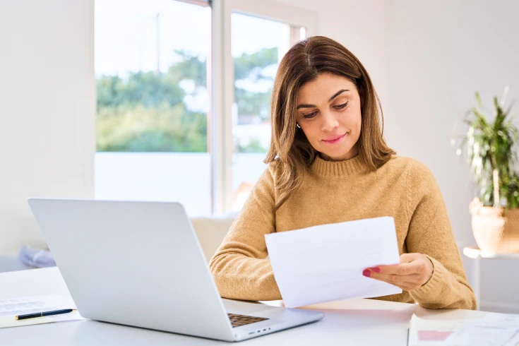 Woman sitting at table with laptop in front of her, reading paper bill