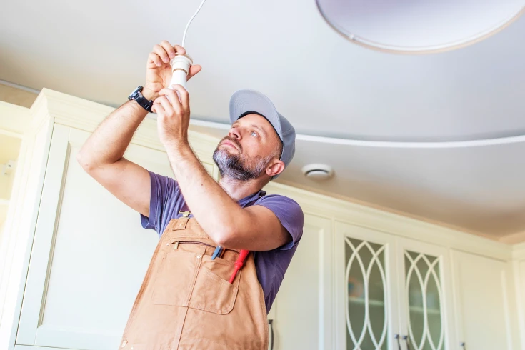 Man in overalls and hat changing hanging a lightbulb in a home