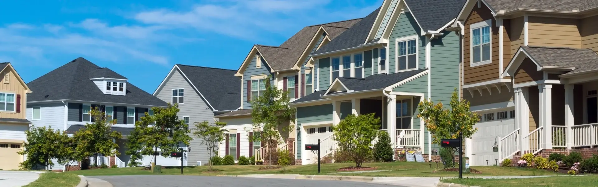 A suburban neighborhood street lined with modern, two‑story single-family homes in various colors, with well-kept lawns, young trees, and a clear blue sky overhead.