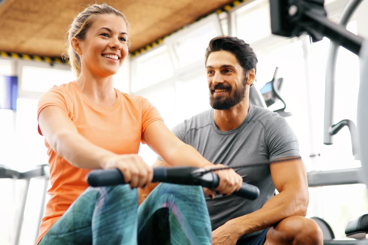 A gym member uses a rowing machine with instruction from a personal trainer