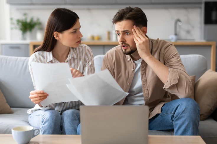 A man and woman are looking at paper while sitting on a couch, the man in shock, wondering why energy prices are going up