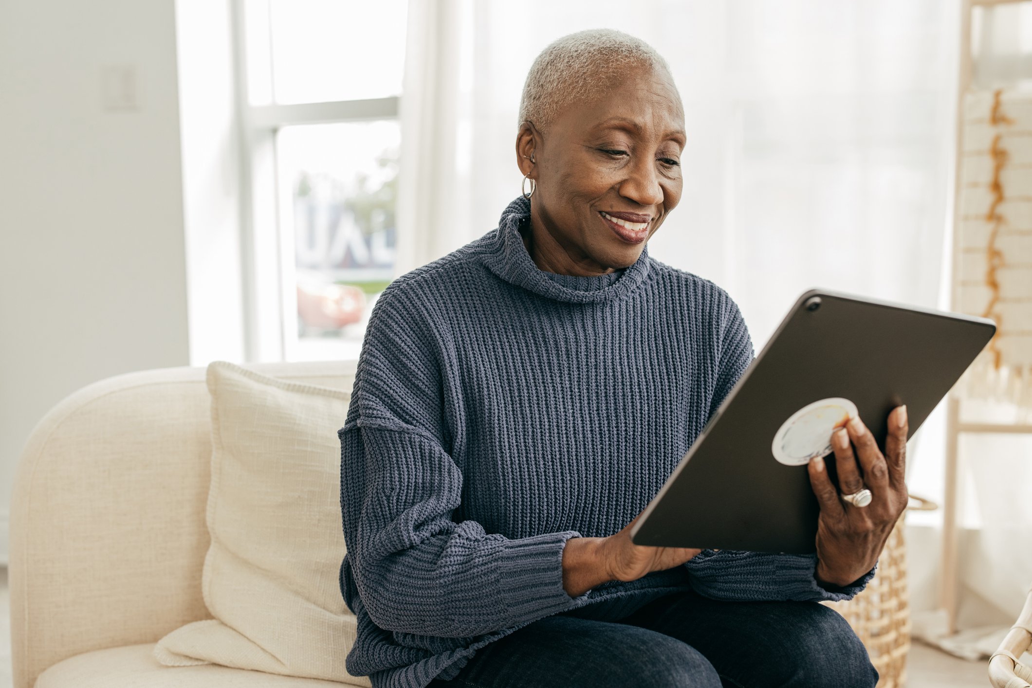 An older woman uses a digital tablet to attend a telehealth appointment while sitting on her couch at home