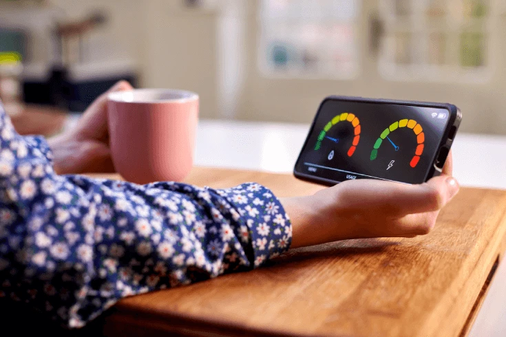 Person sitting at a table, holding a mug and checking their energy usage on a smartphone app.