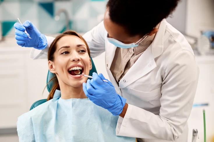 Woman smiling while sitting in the dentist’s chair with the dentist checking her teeth