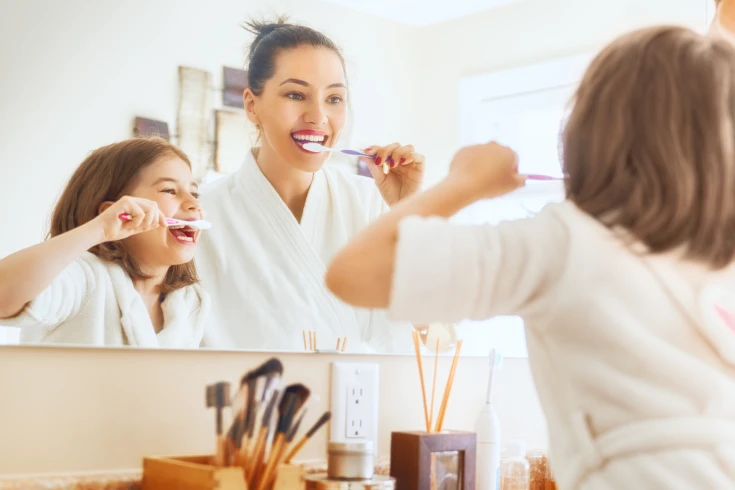 A mother and daughter smile while brushing their teeth