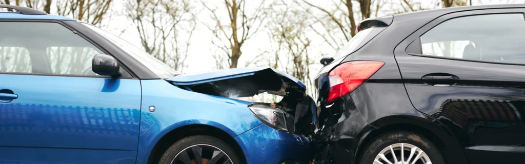 A picture of two cars involved in a crash with a blue car rear-ending a black car. The blue car has a damaged front bumper and the bonnet is also damaged. 
