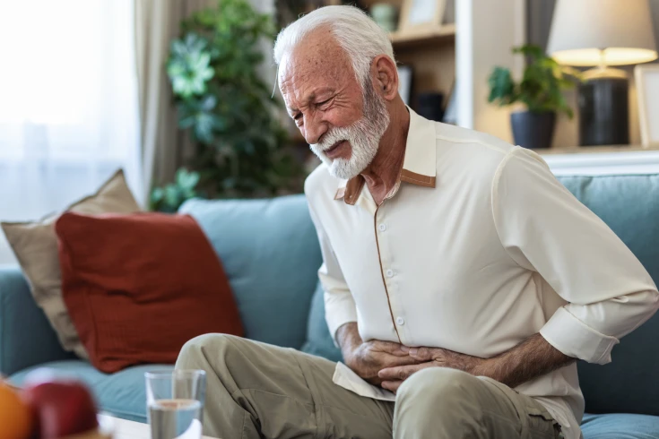 An old man sitting in the couch wincing in pain while holding his stomach with his two hands