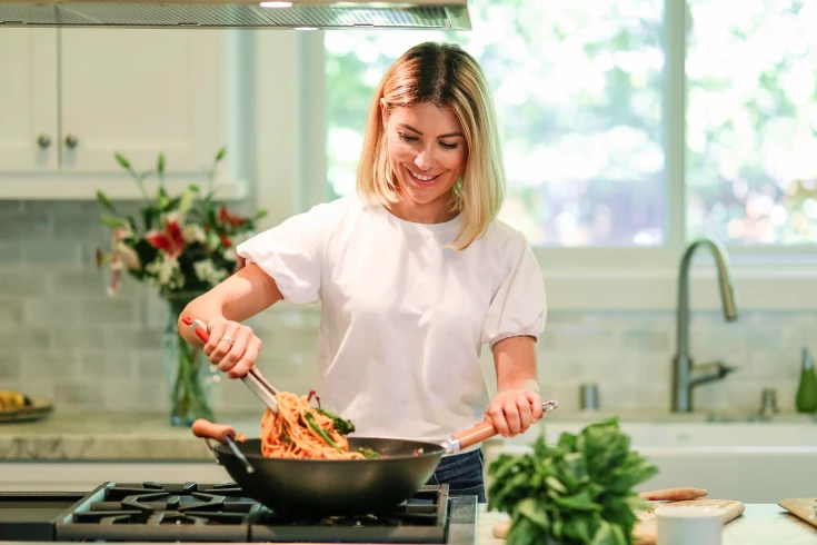Woman tossing noodles in wok on a stovetop in a kitchen