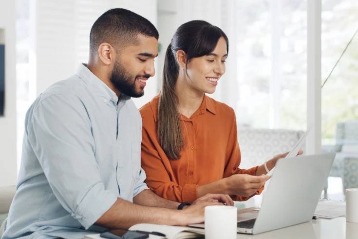 A man and a woman sitting together, man is typing on a laptop, the woman is looking at a paper bill