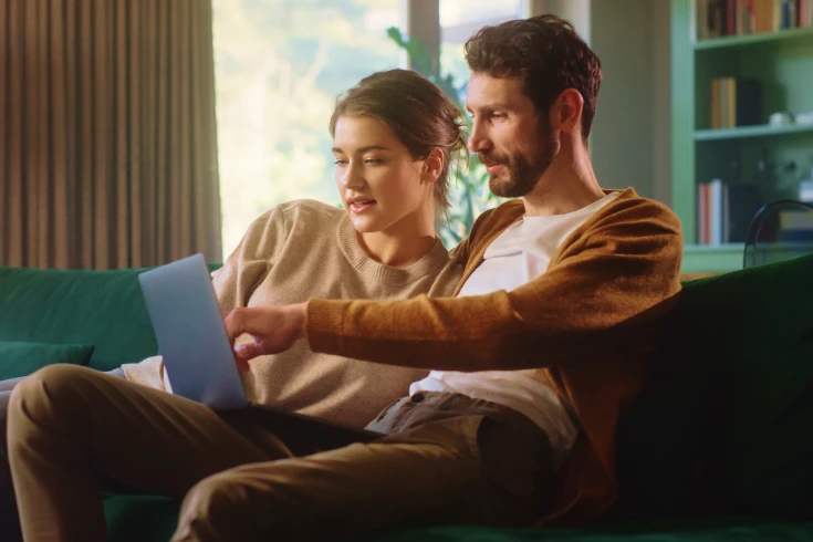 Image of a man and woman sitting on a couch in a living room, looking at a laptop on the man's lap.