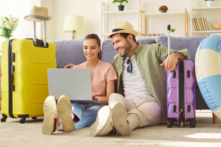 Image of a man and woman sitting on the living room floor with suitcases, both looking at a laptop on the woman's lap.