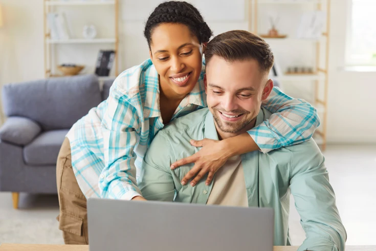 A man and woman looking at a laptop, the man is sitting at a table with the woman's around his shoulder