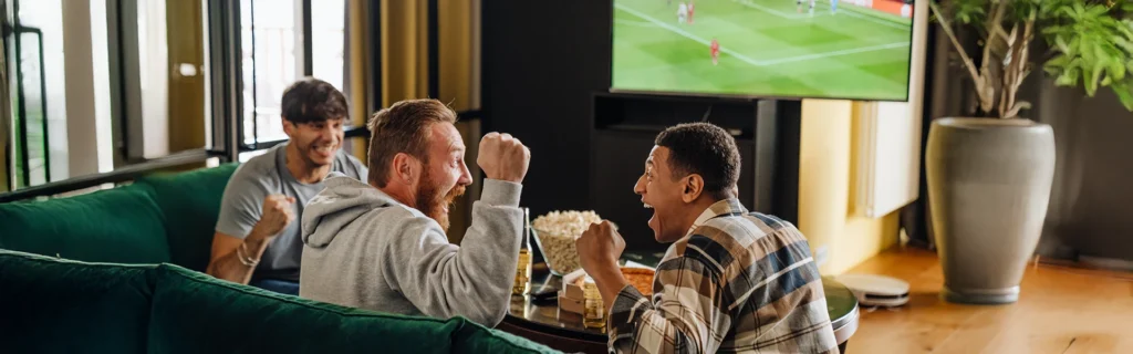 three men sitting on a couch excitedly cheering as they watch a soccer match on TV
