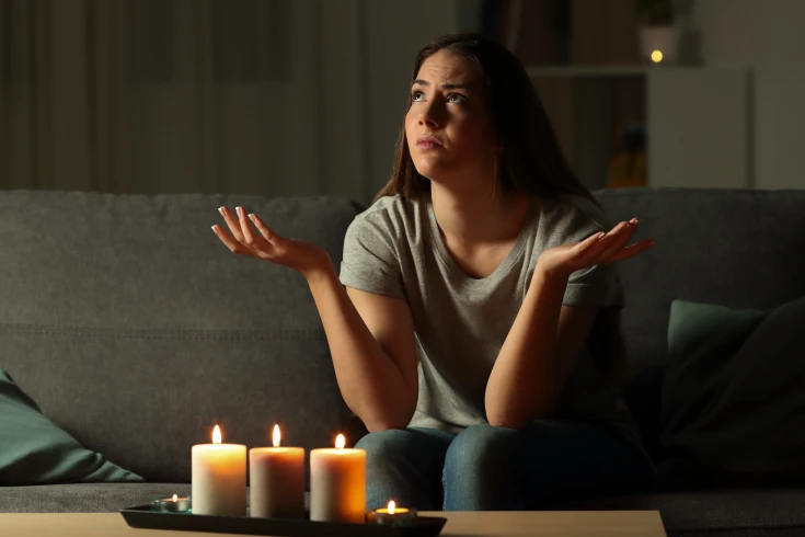 A woman throwing her hands out in annoyance, sitting on a couch in the dark with three candles lit in front of her