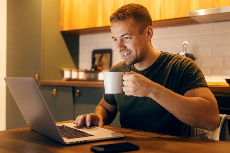 Man sitting at a kitchen table holding a mug and looking at his laptop